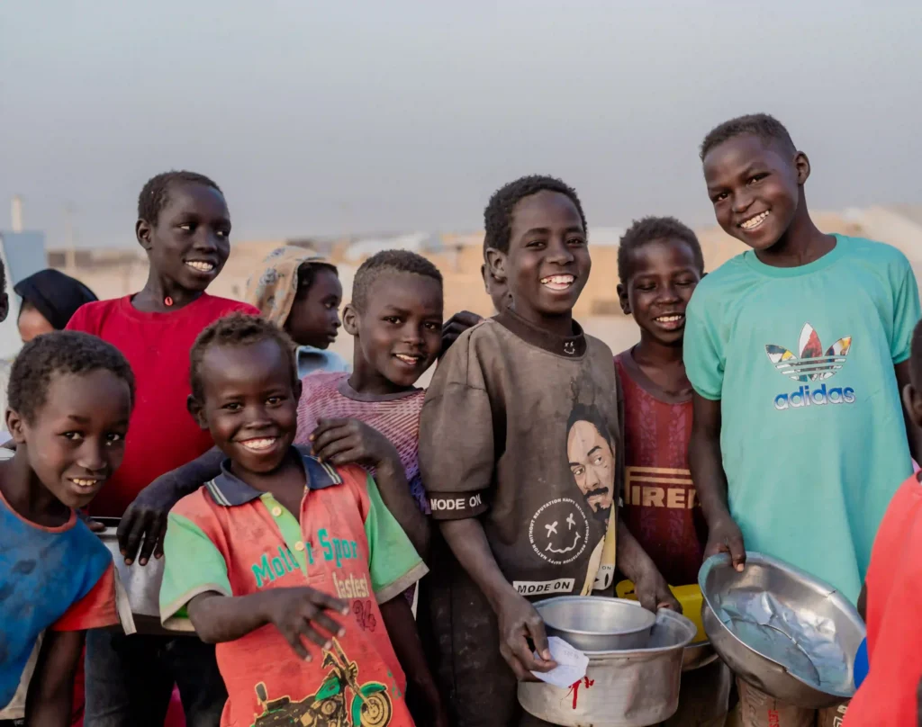A group of Sudanese children and boys smiling and gathered together outdoors in a displacement camp, several holding metal bowls and pots, with tents and shelters visible in the hazy background at dusk.