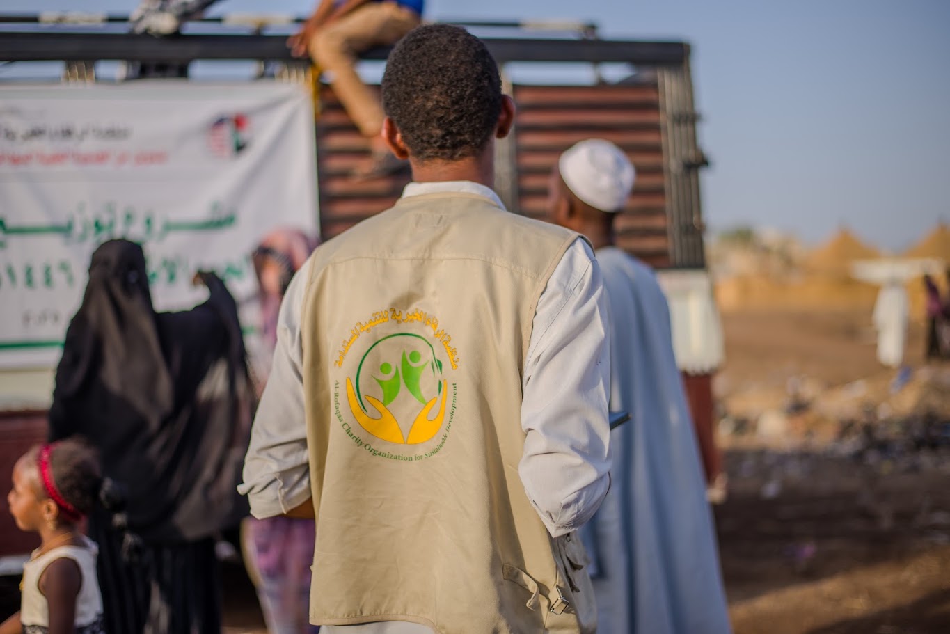 A community outreach worker wearing a beige vest with a humanitarian organization logo stands among people at an outdoor community event, with women and children nearby and a rural settlement visible in the background.