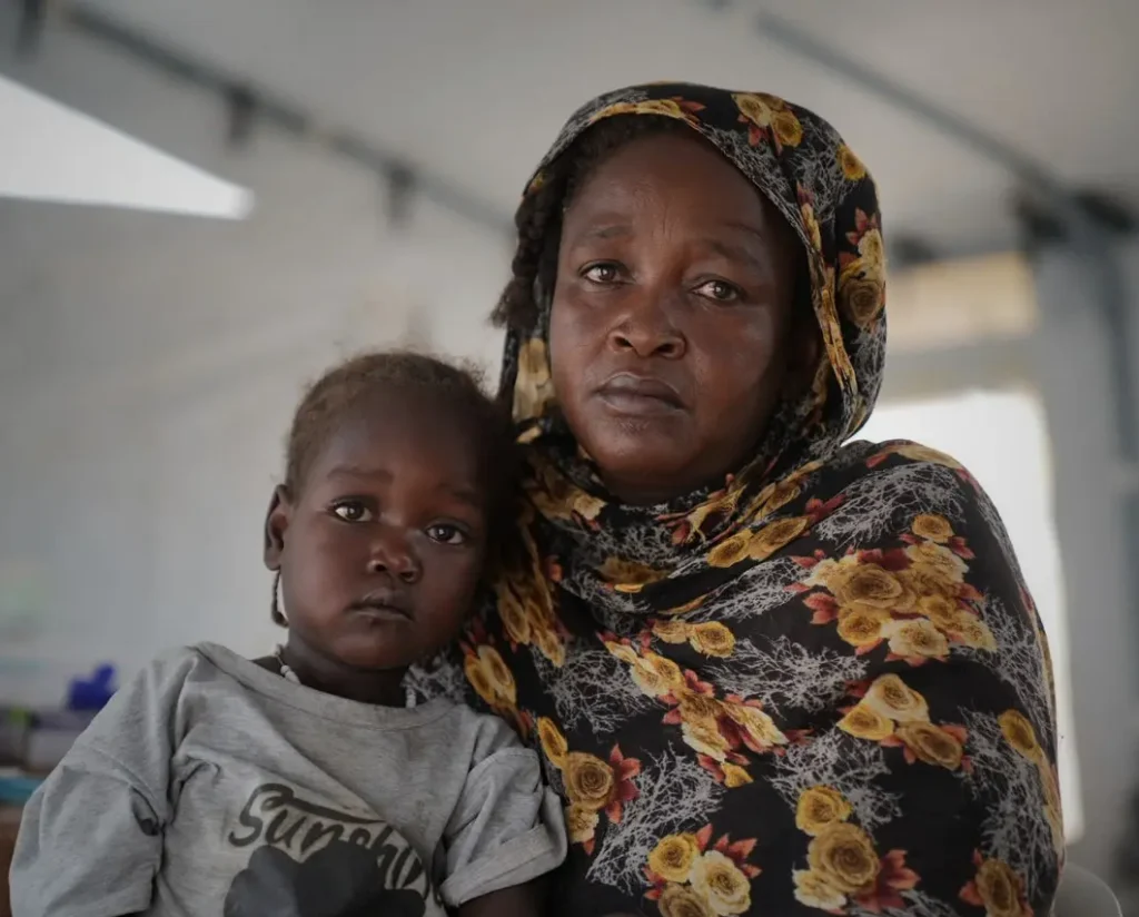 A mother in a floral headscarf holds her young child at a SAMA mobile clinic, both looking directly at the camera.